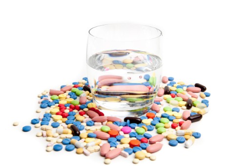 Many prescription drug pills surrounding glass of water